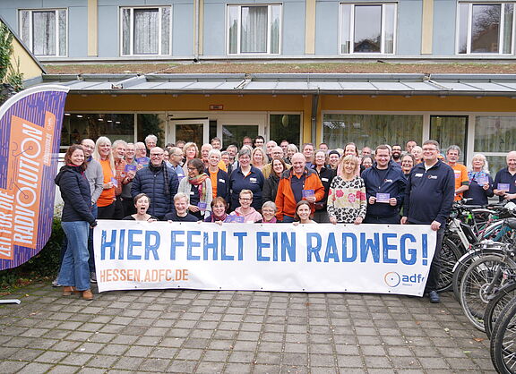 Die Gruppe der ADFC-Aktiven steht vor dem Eingang und hält ein Banner mit der Aufschrift „Hier fehlt ein Radweg“ in die Höhe