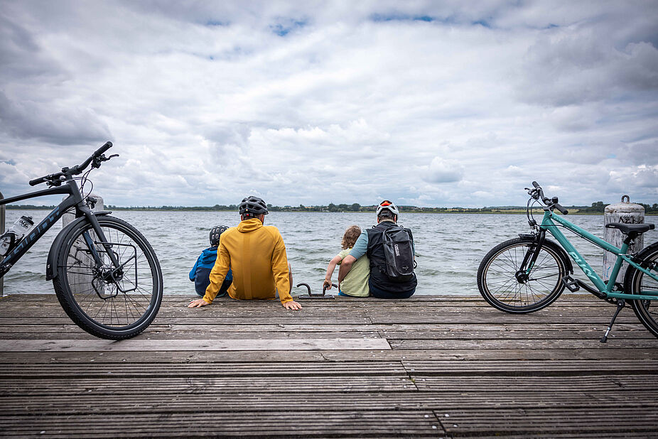 Mit den Kindern auf Fahrradtour Zwei Personen, zwei Kinder auf einem Steg, dahinter Wasser, mit Fahrrädern.