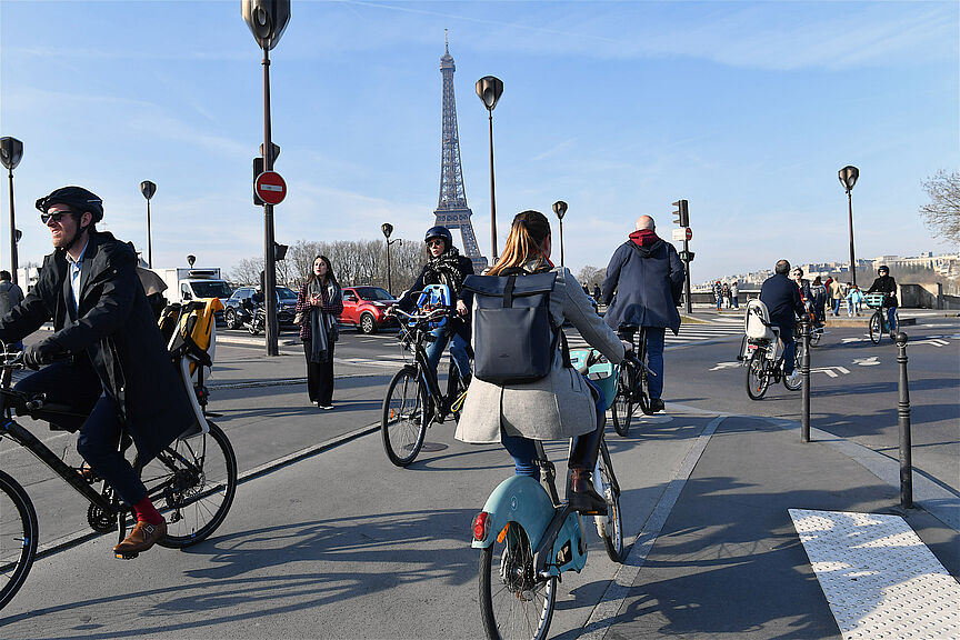 Menschen auf Fahrrädern überqueren eine Straße in Paris. Im Hintergrund ist der Eiffelturm zu sehen.
